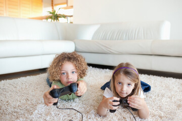 Boy and girl children playing games, lying on shag rug in living room holding wired controllers