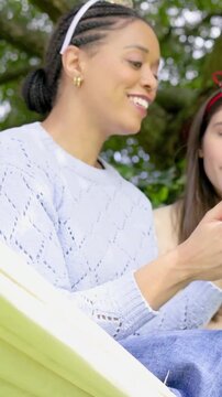 Vertical video: Friend urging woman in blue knit and tiara showing pink phone under tree, smiling