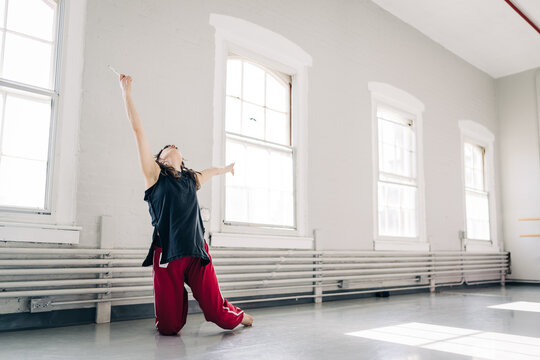 Dancer kneels with arms outstretched in bright, empty studio