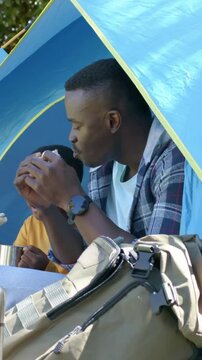 Vertical video: Unwrapping sandwich, dad in plaid shirt feeding son in yellow at blue tent, camping