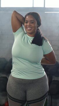 Vertical video: Starting warmup woman in mint top doing triceps stretch in gym by dumbbells