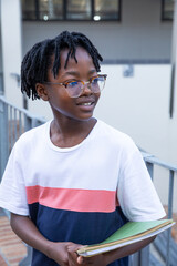 African american teenager standing on outdoor walkway by railing holding green notebooks