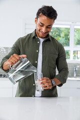 Hispanic man pouring water into tall drinking glass on kitchen island