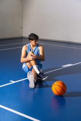 Naklejka premium Asian teenage male basketball player holding knee and stretching leg on court near orange ball