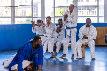 Diverse martial arts students sitting on bench clapping cheering in gis on blue mats in studio