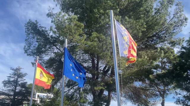 Three flags are hanging from a tree, one of which is a red and yellow flag. The other two flags are blue and white. The scene is peaceful and serene, with the flags fluttering in the breeze
