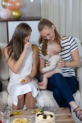 Obraz premium Mother and friend with infant on light-colored sofa in living room sharing gold party horn