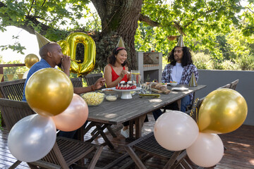Diverse friends sitting at table on deck beside balloons wearing party hats, eating strawberries