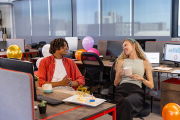 Diverse coworkers celebrating birthday at open-plan office with cake, balloons, coffee mugs