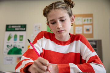 Schoolgirl writing in notebook at classroom desk with pencil by recycling chart