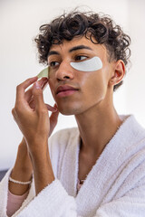 Man wearing bathrobe applying hydrogel eye patches under right eye in grooming area