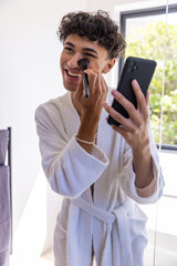 Hispanic man standing at bathroom mirror applying blush with makeup brush, holding smartphone