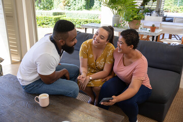 Fototapeta premium Diverse friends sitting on sofa in patio lounge holding smartphone while sipping from pink mug
