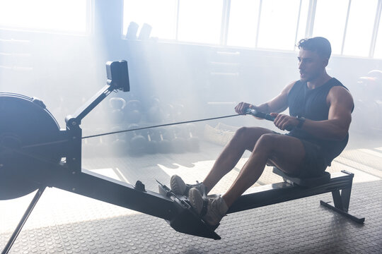 Muscular adult man working on rowing machine near kettlebells in gym under sunlight, copy space