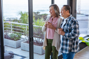 Diverse senior couple standing by sliding doors on balcony holding mugs and watching ocean