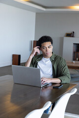 Naklejka premium African american man sitting at dining table using laptop with smartphone in living room