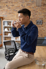 Asian man wearing shirt sitting at desk in office drinking water from glass beside pitcher