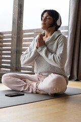 Adult asian woman meditating on yoga mat by glass door with smartphone and headphones