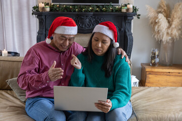 Naklejka premium Asian couple with laptop and pointing at screen wearing santa hats in living room