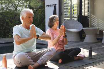 Senior couple meditating while holding hands on yoga mats on wooden poolside deck