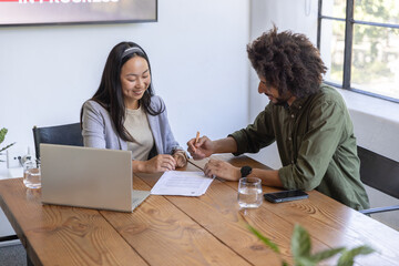 Chinese female colleagues signing printed document with pen at office meeting table, laptop