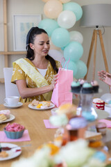 Obraz premium Woman in her thirties opening pink gift bag at wooden dining table with cupcake stand