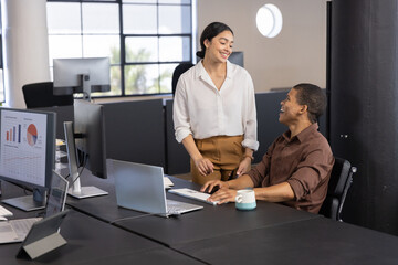 Obraz premium Diverse female coworkers collaborating at desk in office using laptop and charts, coffee mug