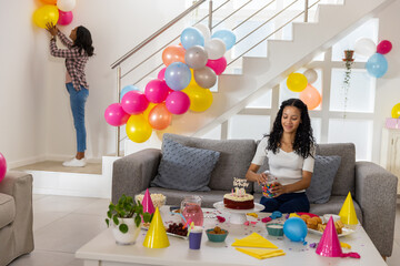 Obraz premium Diverse daughter arranging candy near cake, mother hanging balloons on wall in living room