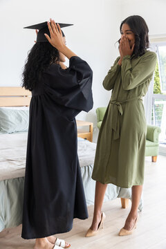 Diverse mom and teenage daughter wearing graduation gown adjusting cap and reacting in shock
