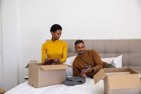 Diverse couple sorting clothing on bed in apartment bedroom with cardboard boxes and smartphone - Powered by Adobe