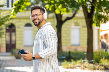 Fototapeta premium Back view of carefree young man tourist in wireless headphones listening favorite energetic disco music dancing outdoors. Caucasian guy traveler walking spinning on urban city street. Town lifestyle.