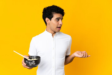 Young Venezuelan man isolated on yellow background with surprise facial expression while holding a bowl of noodles with chopsticks