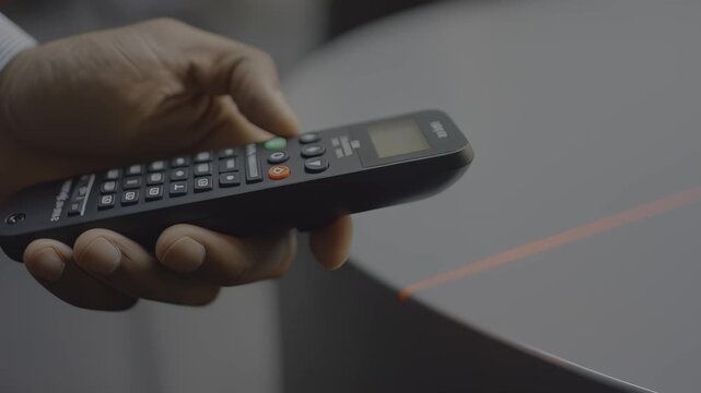 Male hand holding a remote control device, pressing buttons while a red laser beam projects onto a circular surface in a modern indoor setting, hdr video with neutral color