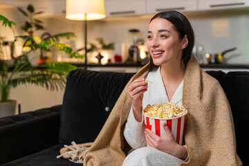 Young woman at night home wears blanket holds popcorn watches comedy smiles talks between bites laughs. Caucasian girl on sofa at evening comments scenes reacts to plot enjoys chill movie time casual.