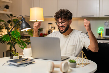 Arabian man at home opens laptop sees approved grant message wins application feels joy. Egyptian guy sitting at table reads good news alert about win and jackpot style reward celebrates success.