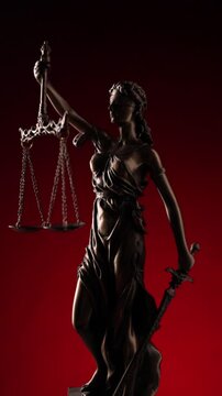 Vertical video of bronze law statue represented by blindfolded goddess spinning and holding a balance signifying impartiality and two broad sources of law on red background.