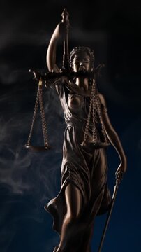 Mysterious smoke swirling around a bronze statue of Lady Justice holding a sword and scales against a dark blue background.