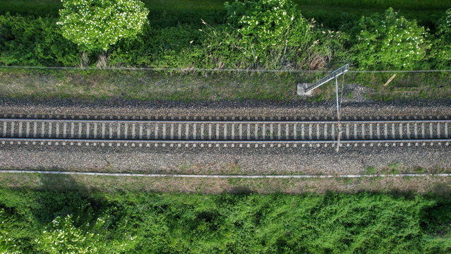 An overhead drone shot captures the parallel lines of a train track cutting through the vibrant green landscape of the Po Valley, Italy