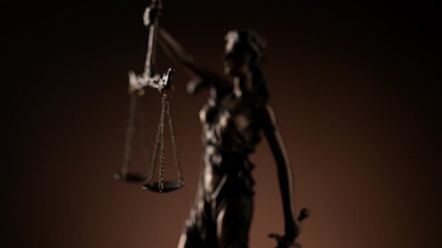 Rack focus shot revealing bronze statue of blindfolded Lady Justice holding scales and sword symbolizing impartiality and legal power on dark brown background.