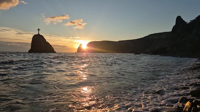 Sunset seascape cliff waves crash on pebble beach with silhouette rock formation and cross structure vertical video