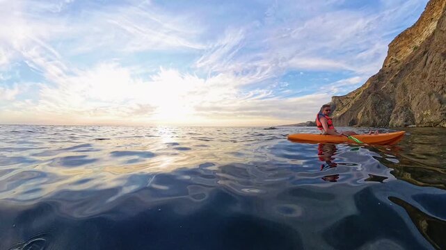 Kayaking sunset ocean man in orange kayak paddles along rocky cliffs during a peaceful golden hour sea adventure on calm water