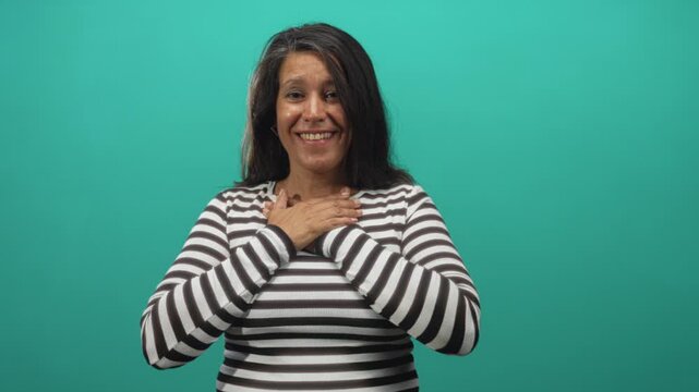 Woman clasping hands to chest in teal studio with striped shirt and gentle smile; gratitude warmth sincerity.