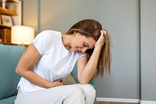 Young woman sitting on sofa at home suffering from severe abdominal pain and stomach ache holding belly in discomfort from menstrual cramps indigestion or gastritis and female health concept