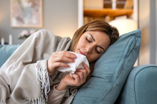 Sick young woman lying on sofa at home blowing nose with paper tissue while suffering from severe cold flu or allergic rhinitis symptoms under a warm blanket healthcare and medical