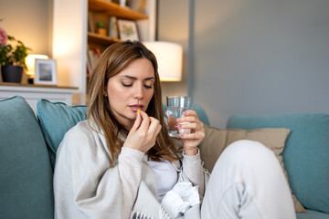 Sick young woman sitting on sofa wrapped in blanket taking a yellow medical pill or vitamin with a glass of water to treat flu symptoms or infection at home healthcare concept