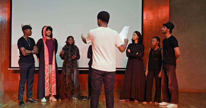 African young adult gen z male man hold scripts paper read play story lines scene standing indoor stage hall. Happy group boy girl clap hand enjoy do work show drama practice day inside campus place