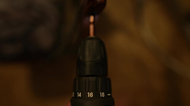 A close-up of a cordless drill/driver being used to drive a screw into a piece of dark, glossy wood. The torque numbers (14, 16, 18) are visible on the drill, and the hand holds it securely. The backg