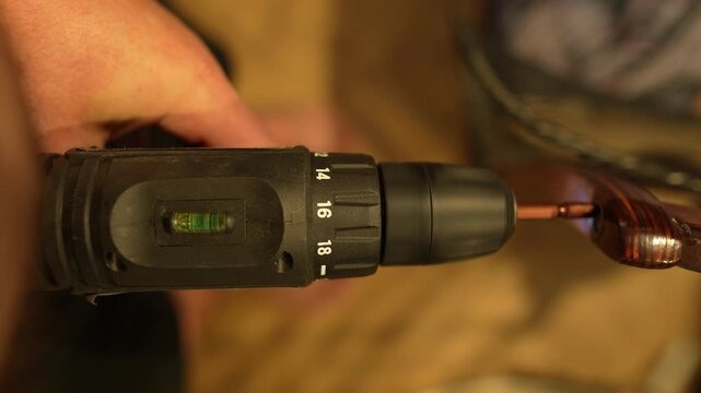A close-up of a cordless drill/driver being used to drive a screw into a piece of dark, glossy wood. The torque numbers (14, 16, 18) are visible on the drill, and the hand holds it securely. The backg