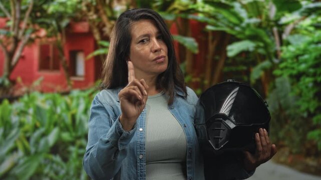 Woman holding a black motorbike helmet and pointing index finger toward camera in front of building courtyard with lush plants; warning resolve.