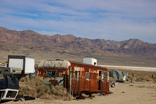Abandoned rusted trailer in a desert scrap yard in Searles Valley, California, USA. Weathered metal, industrial decay and arid mountains under a pale blue sky.